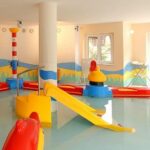 Kinderplanschbecken im Innenbereich mit bunten Spielgeräten, einer kleinen Rutsche und flachem Wasser, umgeben von hellen Wanddekorationen und großen Fenstern.