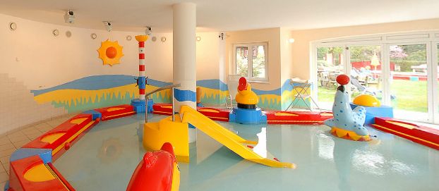 Kinderplanschbecken im Innenbereich mit bunten Spielgeräten, einer kleinen Rutsche und flachem Wasser, umgeben von hellen Wanddekorationen und großen Fenstern.