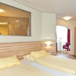 Ein Hotelzimmer mit zwei Einzelbetten mit gelber Bettwäsche, einem hölzernen Kleiderschrank, einem kleinen Tisch und Stuhl am Fenster und einem großen Innenfenster über den Betten.