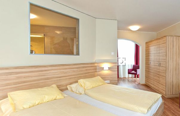 Ein Hotelzimmer mit zwei Einzelbetten mit gelber Bettwäsche, einem hölzernen Kleiderschrank, einem kleinen Tisch und Stuhl am Fenster und einem großen Innenfenster über den Betten.