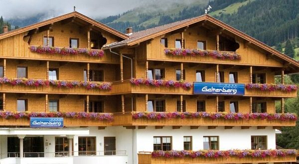 Ein Alpenhotel aus Holz mit mehreren Balkonen, die mit Blumenkästen geschmückt sind, vor einer Kulisse aus grünen Hügeln und Bäumen. Auf den Schildern steht "Galtenberg.