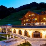 Ein großes Hotel im Chalet-Stil mit Balkonen, Außenpool, Sitzgelegenheiten auf der Terrasse und Bergblick in der Abenddämmerung.