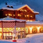 Ein großes Hotel im Chalet-Stil mit warmen Lichtern leuchtet in der Abenddämmerung in einer verschneiten Berglandschaft.