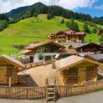 Ein eingezäuntes Gelände mit kleinen Holzhütten liegt zwischen größeren Gebäuden im alpinen Stil und grünen Hügeln unter einem teilweise bewölkten Himmel.