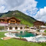 Großes Hotel im Chalet-Stil mit Holzbalkonen, Außenpool, Liegestühlen und Bergkulisse unter teilweise bewölktem Himmel.