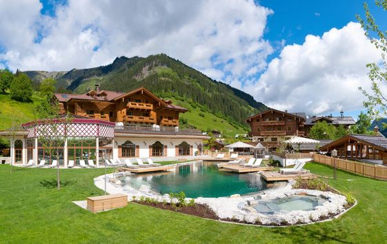 Großes Hotel im Chalet-Stil mit Holzbalkonen, Außenpool, Liegestühlen und Bergkulisse unter teilweise bewölktem Himmel.