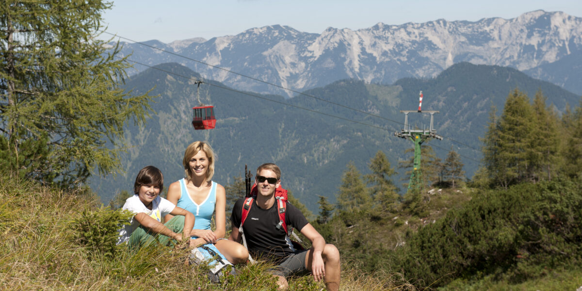 Eine Gruppe von drei Personen sitzt auf einem grasbewachsenen Hügel mit Bergen, Bäumen und einer roten Seilbahn im Hintergrund.