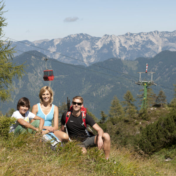 Eine Gruppe von drei Personen sitzt auf einem grasbewachsenen Hügel mit Bergen, Bäumen und einer roten Seilbahn im Hintergrund.