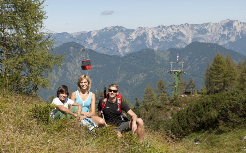 Eine Gruppe von drei Personen sitzt auf einem grasbewachsenen Hügel mit Bergen, Bäumen und einer roten Seilbahn im Hintergrund.