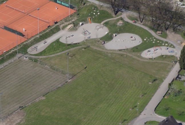Luftaufnahme eines Parks mit einem Skatepark, Spielplatz, Bänken, Tennisplätzen und von Gras und Bäumen gesäumten Wegen.