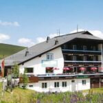 Ein Hotel im Chalet-Stil mit Balkonen und roten Sonnenschirmen auf einer Terrasse, eingebettet in eine grasbewachsene, hügelige Landschaft unter blauem Himmel.