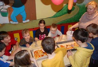 Eine Gruppe von Kindern sitzt um einen Tisch herum und bestreicht den Teig mit Soße, während ein Erwachsener in einem farbenfrohen Raum mit Wandmalereien und Luftballons Pizzen herstellt.