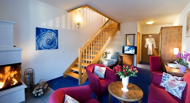 Ein gemütliches Hotelzimmer mit einem Kamin, roten Sesseln, einem Couchtisch mit Blumen, einem Fernseher, einem Schreibtisch, einer Treppe zu einem Loft und einem Bademantel, der am Eingang hängt.