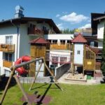 Ein mehrstöckiges Hotel mit Balkonen überblickt einen grasbewachsenen Hof, auf dem sich an sonnigen Tagen ein Spielplatz mit Rutschen, Schaukeln und Klettergerüsten befindet.