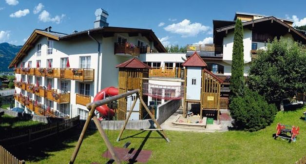 Ein mehrstöckiges Hotel mit Balkonen überblickt einen grasbewachsenen Hof, auf dem sich an sonnigen Tagen ein Spielplatz mit Rutschen, Schaukeln und Klettergerüsten befindet.