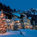 Ein verschneites Hotel mit beleuchteten Weihnachtsbäumen davor, umgeben von Tannenbäumen und Bergen in der Abenddämmerung.