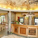 Eine Hotelrezeption mit hölzernem Empfangstresen, Marmorsäulen, Wandmalerei an der Decke und einer Sitzecke mit Sesseln und einem Sofa.