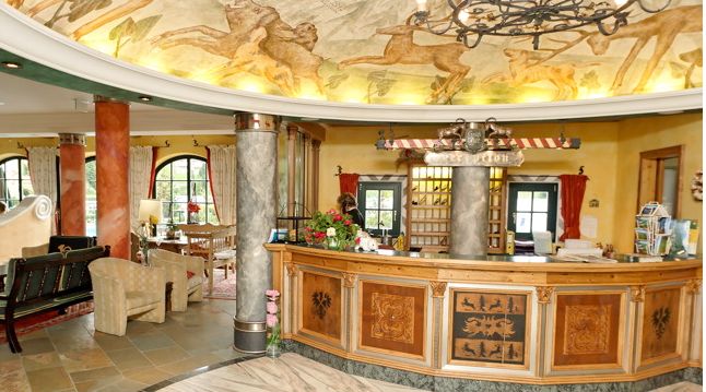 Eine Hotelrezeption mit hölzernem Empfangstresen, Marmorsäulen, Wandmalerei an der Decke und einer Sitzecke mit Sesseln und einem Sofa.