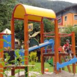 Zwei Kinder spielen auf einem farbenfrohen Spielplatz im Freien mit Rutschen und Klettergerüsten in einer Grünanlage mit Bäumen und nahe gelegenen Gebäuden.