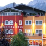 Ein farbenfrohes Hotel mit orangefarbenen und roten Wänden, vielen Fenstern, Balkonen mit Blumen und Außenbeleuchtung, eingebettet in eine bergige Landschaft.