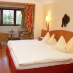 Ein Hotelzimmer mit einem Doppelbett, zwei Stühlen, einem kleinen runden Tisch am Fenster, Holzfußboden und roten und gelben Dekorationsakzenten.