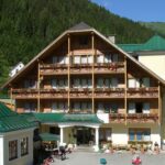 Ein mehrstöckiges Hotel im Chalet-Stil mit Balkonen, Blumenkästen und begrünten Dächern, das an einem sonnigen Tag an einem bewaldeten Berghang liegt.