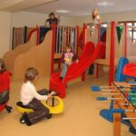 In einem Indoor-Spielzimmer mit Holzspielgeräten können die Kinder auf Rutschen, Fahrgeräten und einem Kickertisch spielen.