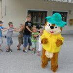 Eine Person in einem Cartoon-Streifenhörnchen-Kostüm führt eine Reihe von Kindern an, die sich vor einem Gebäude aneinander festhalten.