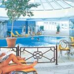 Menschen, die sich auf Liegestühlen an einem Hallenbad mit einer Glaskuppel, Pflanzen und mehreren Personen, die im Becken schwimmen, entspannen.