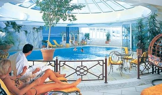 Menschen, die sich auf Liegestühlen an einem Hallenbad mit einer Glaskuppel, Pflanzen und mehreren Personen, die im Becken schwimmen, entspannen.