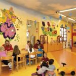 Kinder und Erwachsene sitzen an Tischen und spielen auf dem Boden in einem farbenfroh dekorierten Klassenzimmer mit bunten Wandmalereien und Spielzeug.