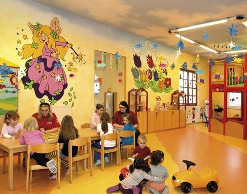 Kinder und Erwachsene sitzen an Tischen und spielen auf dem Boden in einem farbenfroh dekorierten Klassenzimmer mit bunten Wandmalereien und Spielzeug.