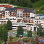 Ein großer, mehrstöckiger Hotelkomplex mit Balkonen, umgeben von Grünanlagen; im Vordergrund sind ein Außenpool, ein Spielplatz und Sitzbereiche zu sehen.