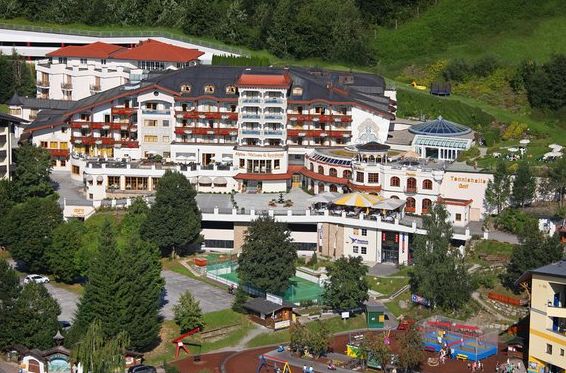 Ein großer, mehrstöckiger Hotelkomplex mit Balkonen, umgeben von Grünanlagen; im Vordergrund sind ein Außenpool, ein Spielplatz und Sitzbereiche zu sehen.