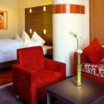 Modernes Hotelzimmer mit einem roten Sessel, einem blumengemusterten Sofa, einem dunklen Couchtisch mit einer Blumenvase und einem Bett mit weißer Bettwäsche im Hintergrund.