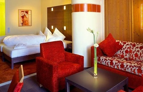 Modernes Hotelzimmer mit einem roten Sessel, einem blumengemusterten Sofa, einem dunklen Couchtisch mit einer Blumenvase und einem Bett mit weißer Bettwäsche im Hintergrund.
