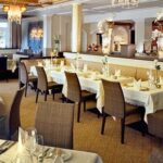 Ein geräumiger, eleganter Restaurantbereich mit ordentlich gedeckten Tischen, gepolsterten Stühlen, Kronleuchtern und dekorativer Beleuchtung.