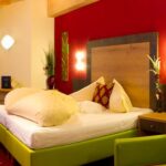 Hotelzimmer mit grünem Bett, weißer Bettwäsche, roter Akzentwand, moderner Einrichtung, zwei Nachttischen, einem Schreibtisch und einem Fenster mit durchsichtigen Vorhängen, die natürliches Licht hereinlassen.