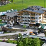 Ein mehrstöckiger Hotelkomplex mit Balkonen, umgeben von grünen Feldern und anderen Gebäuden, mit vor der Tür geparkten Autos und einem Poolbereich auf der linken Seite.