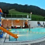 Außenpoolbereich mit Kinderspielplatz, kleiner Wasserrutsche und regenbogenfarbener Rutsche, umgeben von Liegestühlen und Bergen im Hintergrund.