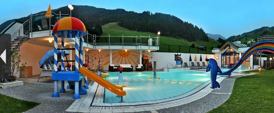 Außenpoolbereich mit Kinderspielplatz, kleiner Wasserrutsche und regenbogenfarbener Rutsche, umgeben von Liegestühlen und Bergen im Hintergrund.