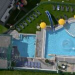 Luftaufnahme eines Freibadkomplexes mit zwei Becken, Liegestühlen, gelben Sonnenschirmen, Wasserrutschen und einer umliegenden grünen Wiese.