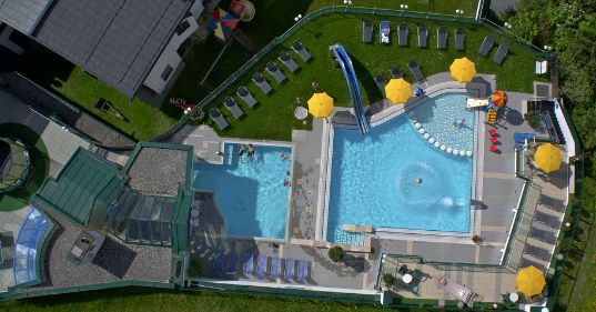 Luftaufnahme eines Freibadkomplexes mit zwei Becken, Liegestühlen, gelben Sonnenschirmen, Wasserrutschen und einer umliegenden grünen Wiese.