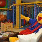 Eine Person in einem farbenfrohen Maskottchenkostüm sitzt auf einem Sofa in einem Indoor-Kinderspielbereich mit Klettergerüsten und einem Ballspielplatz.