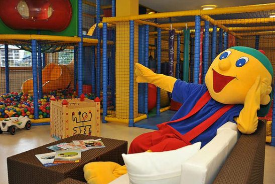 Eine Person in einem farbenfrohen Maskottchenkostüm sitzt auf einem Sofa in einem Indoor-Kinderspielbereich mit Klettergerüsten und einem Ballspielplatz.