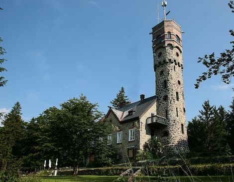 Ein steinernes Gebäude mit einem hohen, runden Aussichtsturm steht zwischen Bäumen unter einem klaren blauen Himmel.