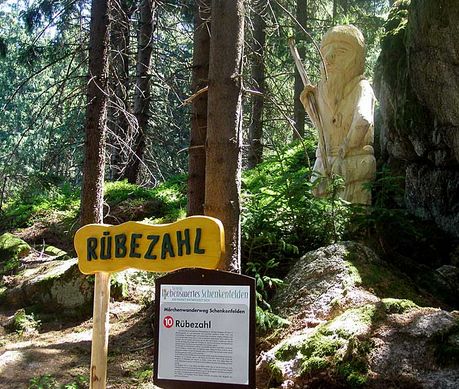 Bewaldetes Gebiet mit einer hölzernen Rübezahl-Statue, die teilweise zwischen Bäumen und Felsen versteckt ist. Ein gelbes Schild und eine Informationstafel mit der Aufschrift "Rübezahl" sind davor zu sehen.