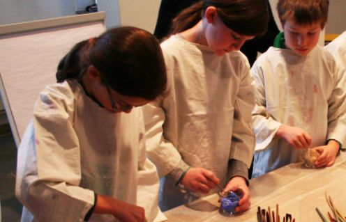 Drei Kinder in weißen Kitteln stehen an einem Tisch und arbeiten mit Malutensilien, insbesondere mit kleinen Behältern und Pinseln.