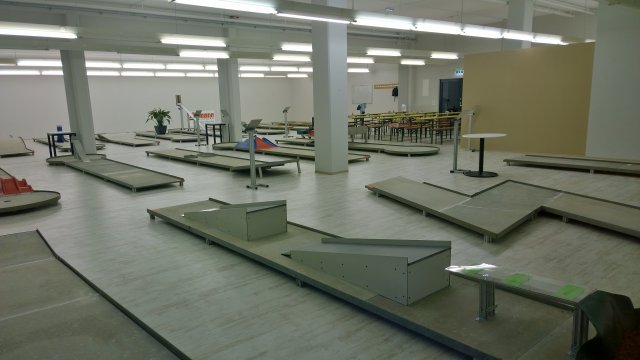 Eine Indoor-Minigolfanlage mit mehreren Bahnen und Hindernissen, angrenzend an einen Sitzbereich mit Tischen und Stühlen unter heller Deckenbeleuchtung.