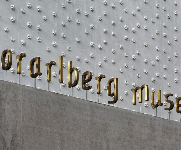 Goldene Buchstaben mit dem Schriftzug "vorarlberg museum" sind auf einer Betonwand mit einer strukturierten, metallischen Oberfläche angebracht, die im Hintergrund erhabene kreisförmige Muster aufweist.